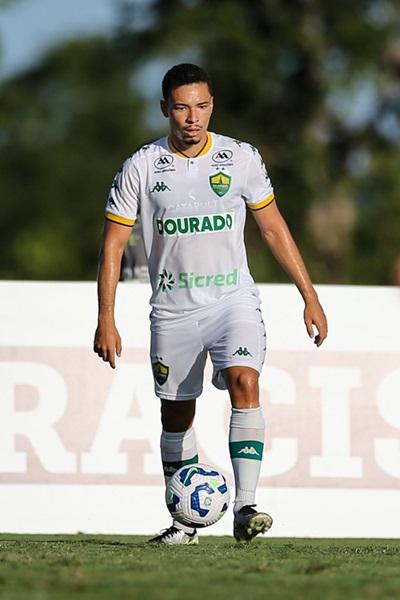 Jogador do Sub-20 do Cuiabá com uniforme branco controlando a bola durante partida, com expressão focada e o sol refletindo no gramado.