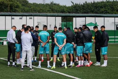 "Grupo de jogadores e comissão técnica do Cuiabá reunidos no centro do campo durante treino, todos usando uniformes de treino verde e preto, com cercas e árvores ao fundo."