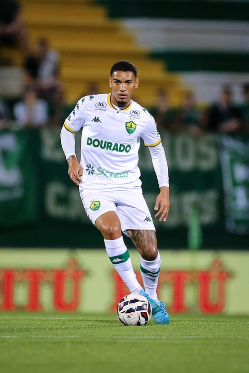 Jogador Denilson, do Cuiabá, conduz a bola durante o jogo contra a Chapecoense, vestindo uniforme branco com detalhes verdes. A partida foi válida pela 8ª rodada da Série B, na Arena Condá.