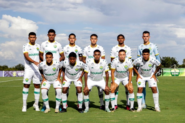 Foto oficial do time Sub-20 do Cuiabá Esporte Clube posado no gramado antes do jogo, com os atletas uniformizados de branco com detalhes verdes e amarelos. Ao fundo, céu azul com nuvens e a faixa do campeonato.