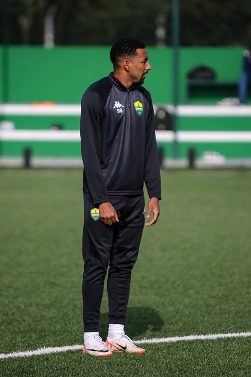Jogador do Cuiabá, usando agasalho preto completo com escudo do clube, durante treino em campo sintético. Ele está de perfil, parado na lateral do gramado, observando o ambiente.