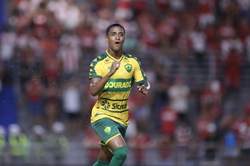 Max comemora seu gol pelo Cuiabá com expressão de alegria, usando o uniforme amarelo e verde, diante da torcida adversária.