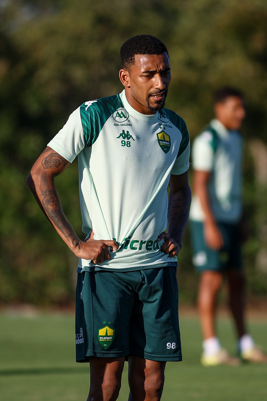 Jogador do Cuiabá em treino ao ar livre, usando uniforme verde-claro e verde-escuro, com as mãos na cintura durante atividade no campo.