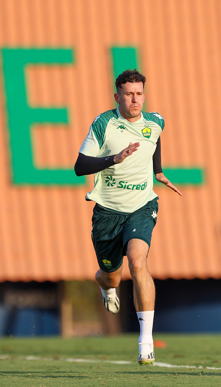 “Jogador do Cuiabá em treino físico, correndo em campo com uniforme de treino verde e branco, fundo desfocado com letras verdes em painel laranja.”