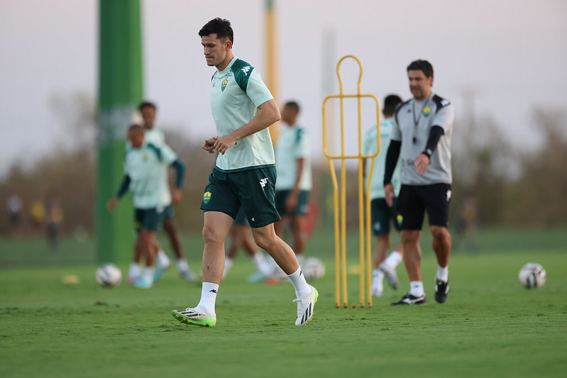 “Jogador do Cuiabá participando de atividade com bola no campo de treino, cones e boneco de treinamento ao fundo, acompanhado pela comissão técnica.”