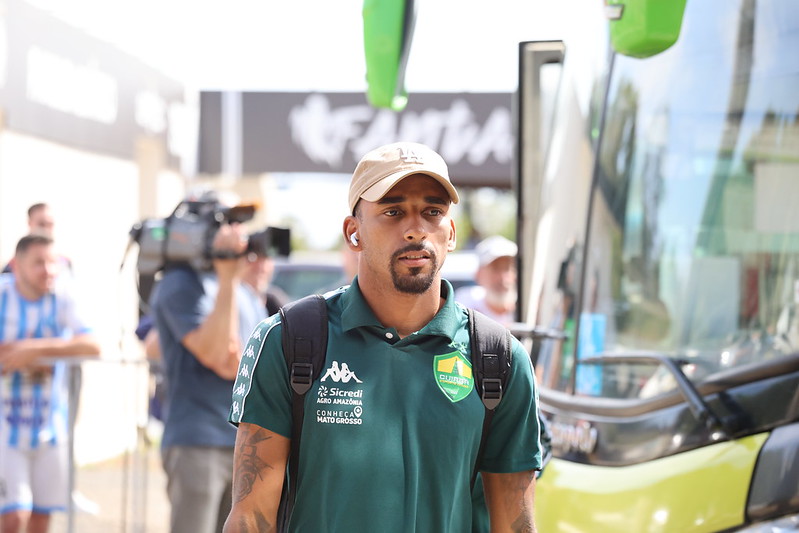 Atleta do Cuiabá chegando para o jogo, vestindo camisa polo verde do clube, boné bege e mochila, em frente ao ônibus da equipe.