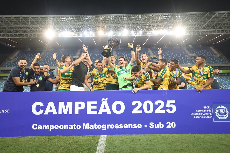 Jogadores do Cuiabá Sub-20 erguendo a taça de campeão 2025 do Campeonato Mato-grossense, sorrindo e comemorando atrás de uma faixa azul que destaca o título, dentro da Arena Pantanal iluminada.