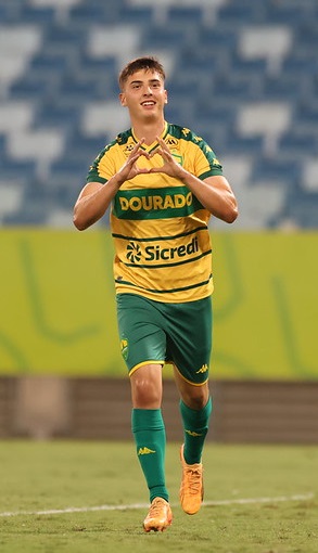 Atacante do Cuiabá Sub-20, com uniforme amarelo e verde, comemorando gol com gesto de coração feito com as mãos, em campo na Arena Pantanal.