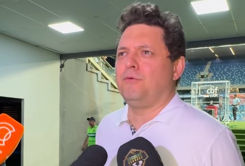 Entrevista em zona mista de estádio de futebol, com homem de camisa polo branca falando à imprensa. Microfones de emissoras de TV e escudo de clube de futebol aparecem em destaque, arquibancadas ao fundo.