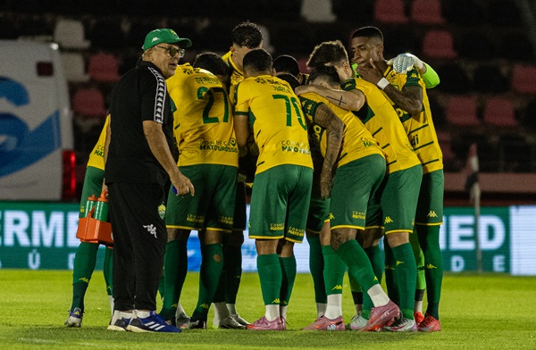 Um grupo de jogadores de futebol com uniformes amarelos e verdes, e um treinador, se reúnem em campo, possivelmente durante uma discussão tática ou celebração. O treinador, um homem mais velho com óculos e boné, está à esquerda, olhando para os jogadores que estão aglomerados no centro.