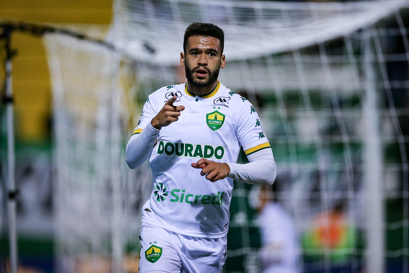 "Patrick de Lucca, volante do Cuiabá, celebra após marcar, apontando com a mão direita. Ele usa o uniforme de jogo branco, em frente a uma trave com rede no fundo."