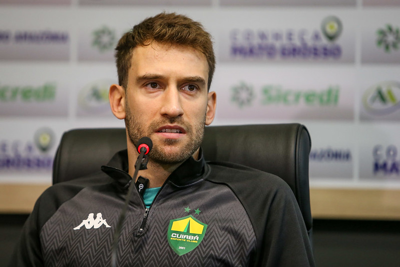 "Alan Empereur, zagueiro e capitão do Cuiabá, em coletiva de imprensa usando agasalho oficial do clube e falando ao microfone."