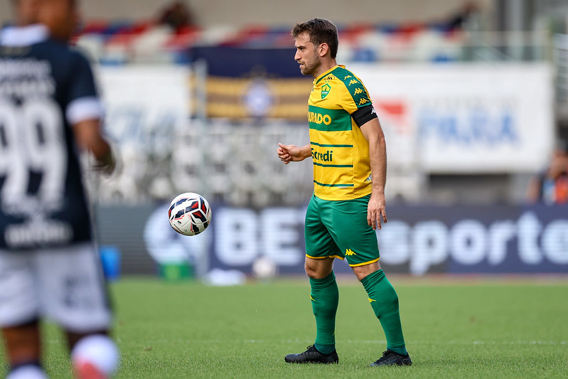 Alan Empereur, um jogador de futebol do Cuiabá, está no campo em seu uniforme verde e amarelo. Ele olha para uma bola de futebol que está no ar, perto dele. Há outro jogador com uma camisa escura no lado esquerdo da imagem, ligeiramente fora de foco.