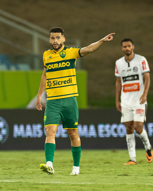 O jogador Patrick de Lucca do Cuiabá, vestindo a camisa branca com detalhes em verde e amarelo, com os patrocinadores 'Dourado' e 'Sicredi' em destaque, comemora um gol apontando para frente em um campo de futebol à noite.