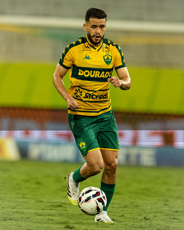 O jogador Patrick de Lucca do Cuiabá, vestindo a camisa branca com detalhes em verde e amarelo, com os patrocinadores 'Dourado' e 'Sicredi' em destaque, comemora um gol apontando para frente em um campo de futebol à noite.