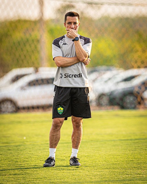 O técnico do Cuiabá Esporte Clube, Eduardo Barros, em pé no gramado do campo de treinamento, com os braços cruzados e a mão direita apoiada no queixo, em pose pensativa. Ele veste o uniforme de treino do clube, composto por uma camisa cinza com o patrocínio Sicredi e shorts pretos.
