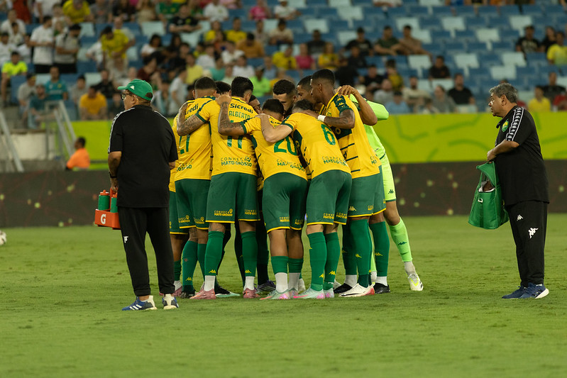 Um grupo de jogadores de futebol com uniformes amarelos e verdes, e um treinador, se reúnem em campo, possivelmente durante uma discussão tática ou celebração. O treinador, um homem mais velho com óculos e boné, está à esquerda, olhando para os jogadores que estão aglomerados no centro.