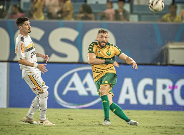 O Lateral Sander com a camisa amarela e verde do Cuiabá chuta a bola com o pé direito, em uma partida anrena pantanal. O marcador do novorizontino, vestindo camisa branca com detalhes pretos e amarelos e calção branco, observa a jogada ao lado. O campo é de grama, e a arquibancada azul e branca aparece desfocada ao fundo.