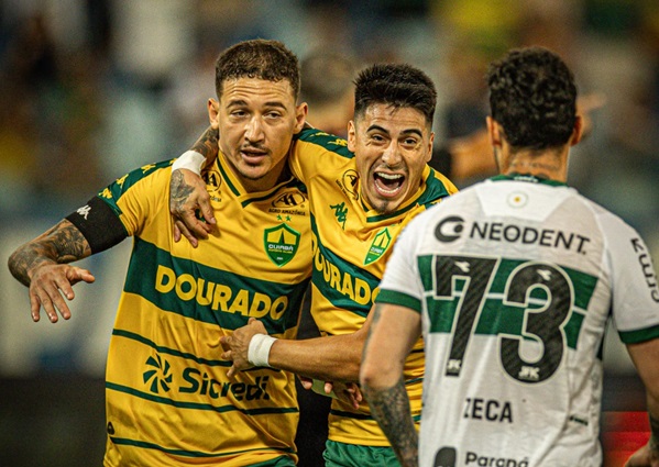 Dois jogadores do Cuiabá, em uniformes amarelos e verdes, se abraçam e comemoram um gol com expressões de alegria intensa. Um jogador adversário, em uniforme branco e verde com o número 73 nas costas, observa ao lado direito.