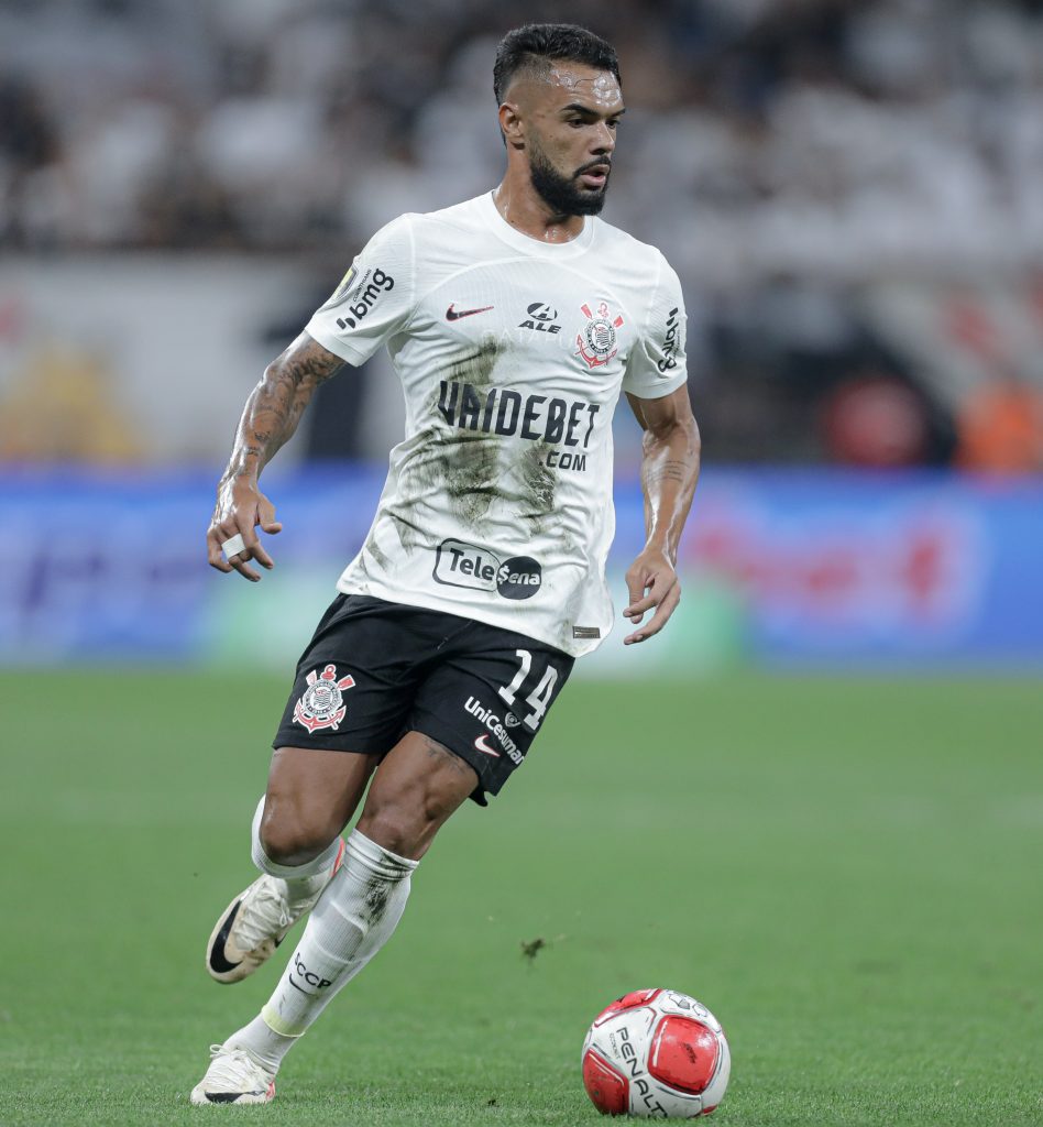 Volante Raniele do Corinthians, vestindo o uniforme branco manchado de lama e suor, está correndo em campo com a bola nos pés. Ele tem tatuagens nos braços e usa o número 14. O jogador está olhando para o lado direito, concentrado na jogada.