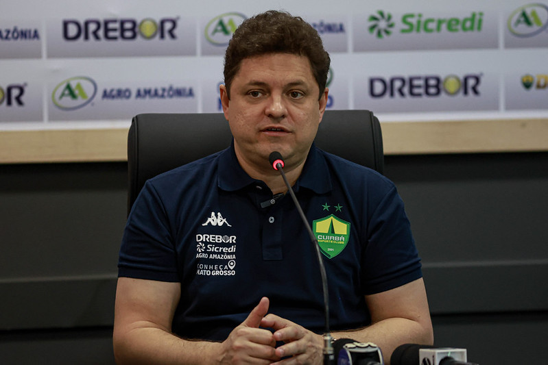 presidente Cristiano Dresch do Cuiabá fala em coletiva de imprensa, sentado à mesa com microfone, usando camisa polo do clube e painel com patrocinadores ao fundo.