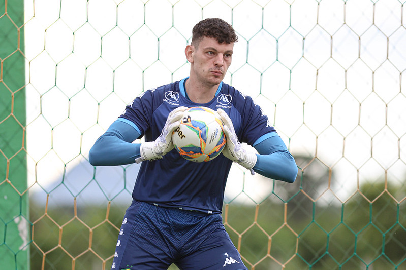 O goleiro, Arthur Bittencourt, em campo, olhando para a direita e segurando uma bola de futebol. Ele usa luvas brancas e uniforme azul-petróleo. O goleiro está entre as traves do gol e uma rede verde pode ser vista atrás dele.