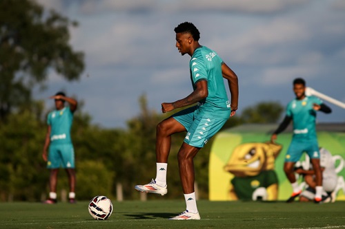 Guilherme Mariano durante treino do Cuiabá, com uniforme verde, controlando a bola no gramado sob o sol, enquanto outros jogadores aparecem ao fundo no centro de treinamento.