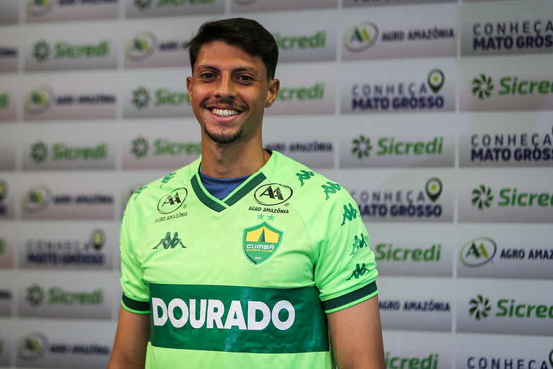 Goleiro Guilherme Nogueira, sorrindo e olhando diretamente para a câmera. Ele veste uma camisa de futebol verde-limão e verde, com a palavra "DOURADO" em letras grandes na parte inferior. Ele está posando na frente de um painel de patrocinadores com logotipos como Sicredi e Agro Amazônia, com a frase "CONHEÇA MATO GROSSO" visível.