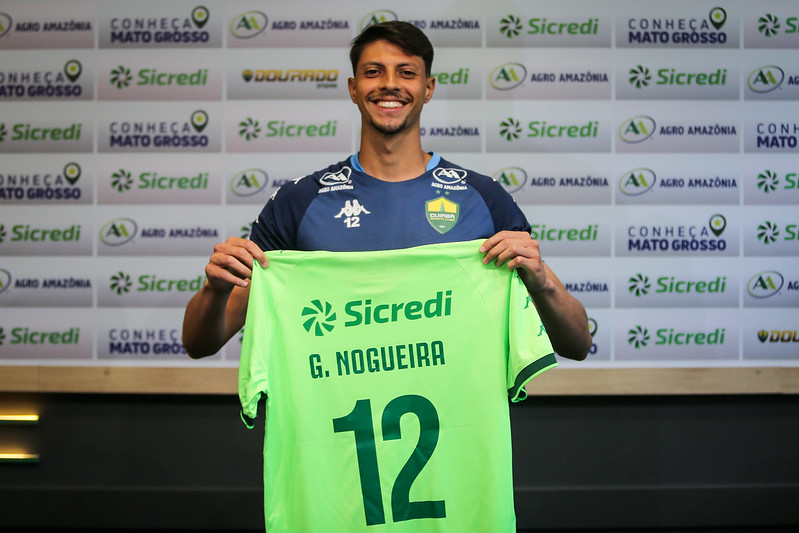 O goleiro Guilherme Nogueira sorrindo e segurando uma camisa verde-clara com o nome 'G. NOGUEIRA' e o número '12' em verde escuro, sobre um fundo com logotipos de patrocinadores.