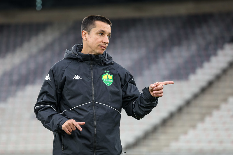 Técnico Eduardo Barros, com um agasalho preto do Cuiabá, em um estádio, gesticulando com a mão direita e apontando para frente ou para o lado, possivelmente dando instruções.