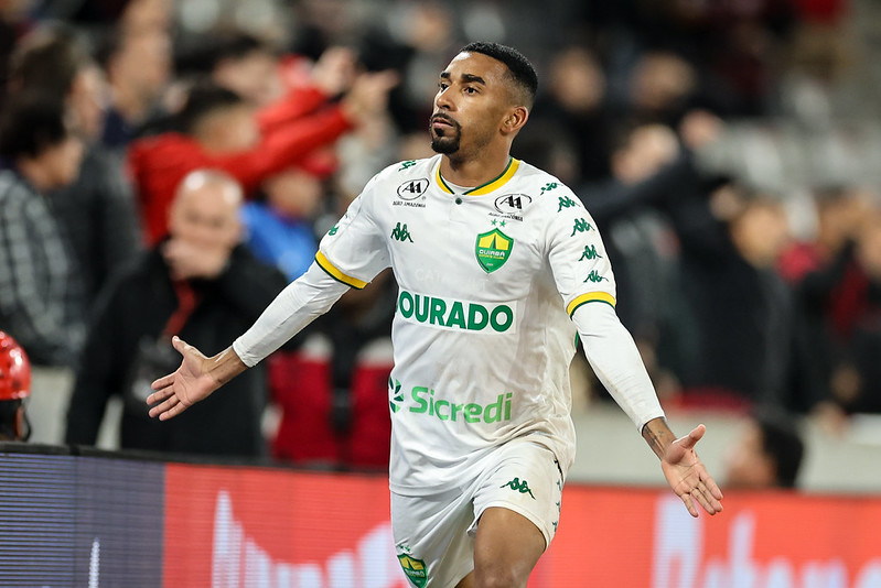 O lateral Matheusinho, do Cuiabá, celebrando em campo, vestindo a camisa branca do time. Ele está com os braços abertos e olhando para cima, em frente à arquibancada com torcedores ao fundo.