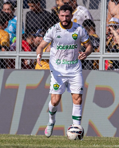 Sander, jogador do Cuiabá, conduz a bola durante partida oficial. Ele veste o uniforme branco do clube, com detalhes verdes e o escudo do Dourado no peito, enquanto o público aparece ao fundo nas arquibancadas.