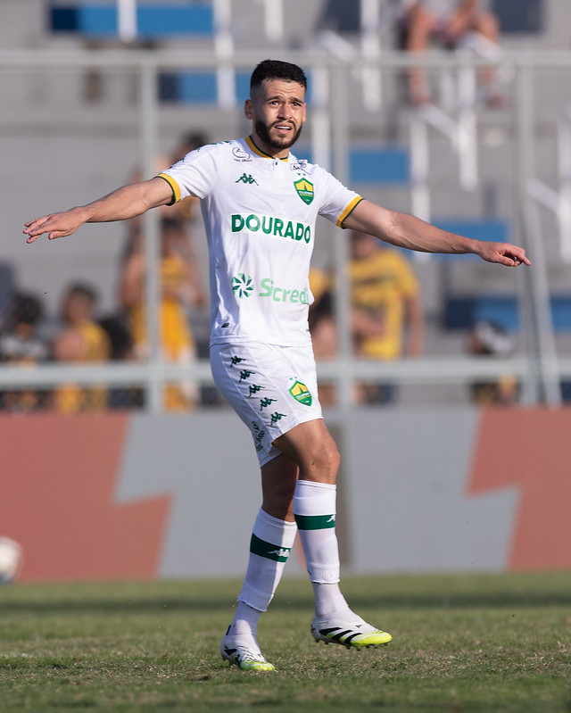 Patrick de Lucca em campo, com os braços abertos, usando o uniforme branco de jogo do time (Dourado).