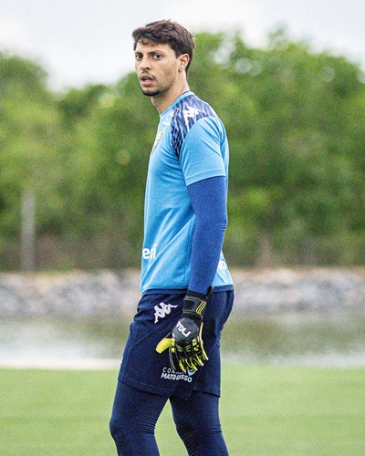 O goleiro Guilherme Nogueira, em um plano médio, usando uniforme de goleiro azul-claro com mangas longas azuis-escuras e luvas pretas e amarelas. Ele está no campo de treinamento, olhando para o lado.