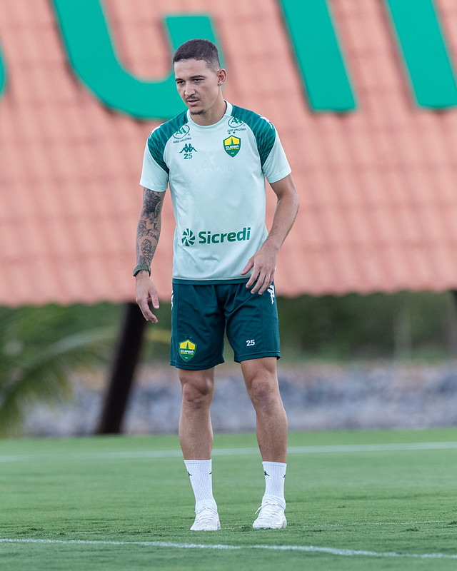 O jogador Alison Safira (identificado como Deyverson) em pé no campo de treino. Ele veste uma camisa de treino clara com mangas verde-escuras e um calção escuro com o número 25. Ele está olhando para baixo com uma expressão calma. O fundo é desfocado com tons de verde e laranja.