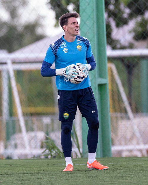 Goleiro Luan Polli em campo, usando uniforme de treino azul e escuro. Ele está segurando uma bola de futebol com luvas brancas.