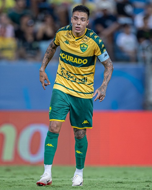 Carlos Alberto em campo, vestindo o uniforme amarelo e verde do clube. Ele está com os braços tatuados expostos e olhando para a frente.