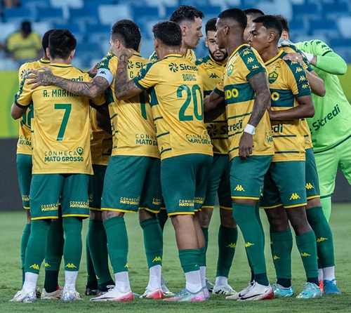 Um grupo de jogadores de futebol com uniformes amarelos e verdes, e um treinador, se reúnem em campo, possivelmente durante uma discussão tática ou celebração. O treinador, um homem mais velho com óculos e boné, está à esquerda, olhando para os jogadores que estão aglomerados no centro.