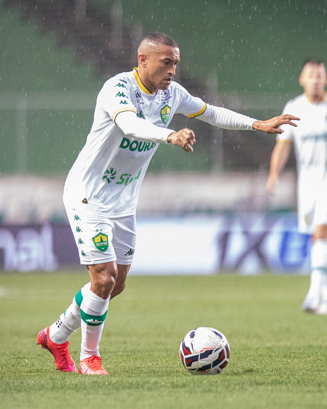 Nino Paraíba com a bola no pé durante um jogo, vestindo o uniforme branco de mangas longas e estendendo os braços.