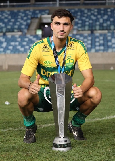 O jogador Renan Carrasco agachado no gramado, sorrindo e segurando um troféu grande e prateado com as duas mãos. Ele veste o uniforme do Cuiabá e faz um sinal de positivo (joia) com a mão esquerda, com a arquibancada azul ao fundo.