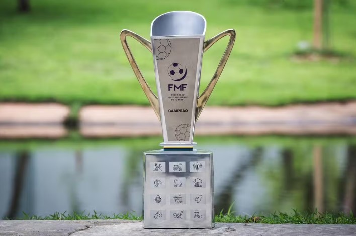 Troféu oficial do Campeonato Mato-Grossense da FMF exibido ao ar livre, com design moderno em tons prateados e dourados, apoiado sobre base decorada com ícones representando elementos do estado de Mato Grosso.