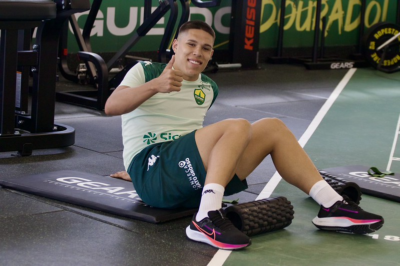 O jogador de futebol Caleb, do Cuiabá EC, está sentado em um colchonete preto de academia, sorrindo e fazendo um sinal de "positivo" com o polegar. Ele veste o uniforme de treino do clube, composto por uma camiseta verde-clara com detalhes escuros e shorts verde-escuro com a inscrição "Conheça Mato Grosso". O atleta calça tênis esportivos pretos com detalhes em rosa e branco. Ao seu lado, no chão de uma academia moderna, há um rolo de liberação miofascial e equipamentos de musculação ao fundo.