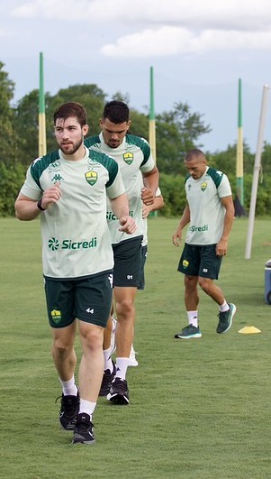 Três jogadores de futebol em primeiro plano realizam um treino de corrida leve no gramado, desta vez calçando tênis esportivos. Eles usam o uniforme oficial de treino com o patrocínio da Sicredi. Ao fundo, membros da comissão técnica organizam equipamentos próximos a uma baliza de treino e uma bandeira amarela de escanteio aparece no canto inferior direito.