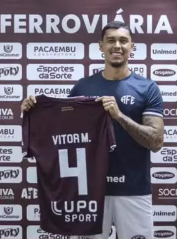 O zagueiro Vitor Mendes sorri durante sua apresentação oficial na Ferroviária. Ele segura a camisa grená do time, que exibe seu nome "VITOR M.0" e o número 4.