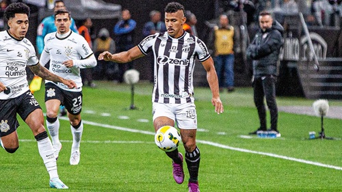 O jogador Fernando Sobral, vestindo o uniforme listrado em preto e branco do Ceará SC, controla a bola com o pé direito durante uma partida em um gramado molhado. Ele está cercado por dois jogadores do Corinthians, que vestem uniformes brancos. Ao fundo, vê-se a área técnica com membros da comissão e arquibancadas desfocadas.