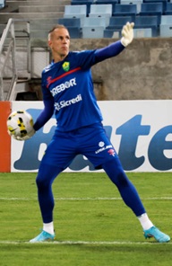 Fotografia de corpo inteiro do goleiro João Carlos em campo durante um jogo ou treino. Ele veste o uniforme azul de goleiro do Cuiabá, composto por camisa azul com detalhes vermelhos e o patrocínio "Drebor" e "Sicredi", além de calças térmicas e shorts azuis. Ele segura uma bola de futebol com a mão direita e mantém o braço esquerdo estendido, gesticulando para orientar o time. Ao fundo, veem-se as arquibancadas vazias de um estádio e placas de publicidade.
