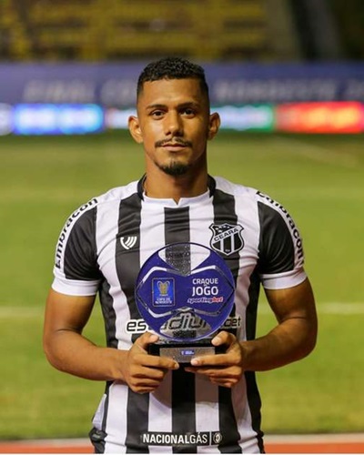 etrato de meio corpo do jogador Fernando Sobral segurando o troféu de "Craque do Jogo" da Copa do Nordeste. Ele veste a camisa oficial do Ceará SC, com listras verticais pretas e brancas e o escudo do clube no peito. O jogador olha diretamente para a câmera com um leve sorriso. O fundo mostra o gramado e as luzes de um estádio à noite.