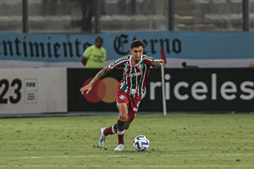 O zagueiro Vitor Mendes, vestindo o uniforme tricolor do Fluminense, conduz a bola com o pé direito durante uma partida de futebol no estádio.