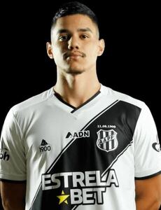 Retrato frontal do zagueiro Edson Rogério posando com o uniforme oficial da Ponte Preta. Ele veste uma camisa branca com uma faixa diagonal preta, onde se vê o escudo do clube (AAPP) e o patrocínio "Estrela Bet". O fundo é inteiramente preto, destacando o rosto e o uniforme do atleta.