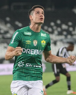 O jogador Matheus Barbosa comemora vibrando intensamente durante uma partida pelo Cuiabá. Ele veste a camisa verde do clube com detalhes em amarelo e o patrocínio "Drebor". O jogador está com os punhos cerrados, braços flexionados e cabeça erguida, expressando euforia. A foto foi tirada em um estádio à noite, com as luzes e o gramado visíveis ao fundo.